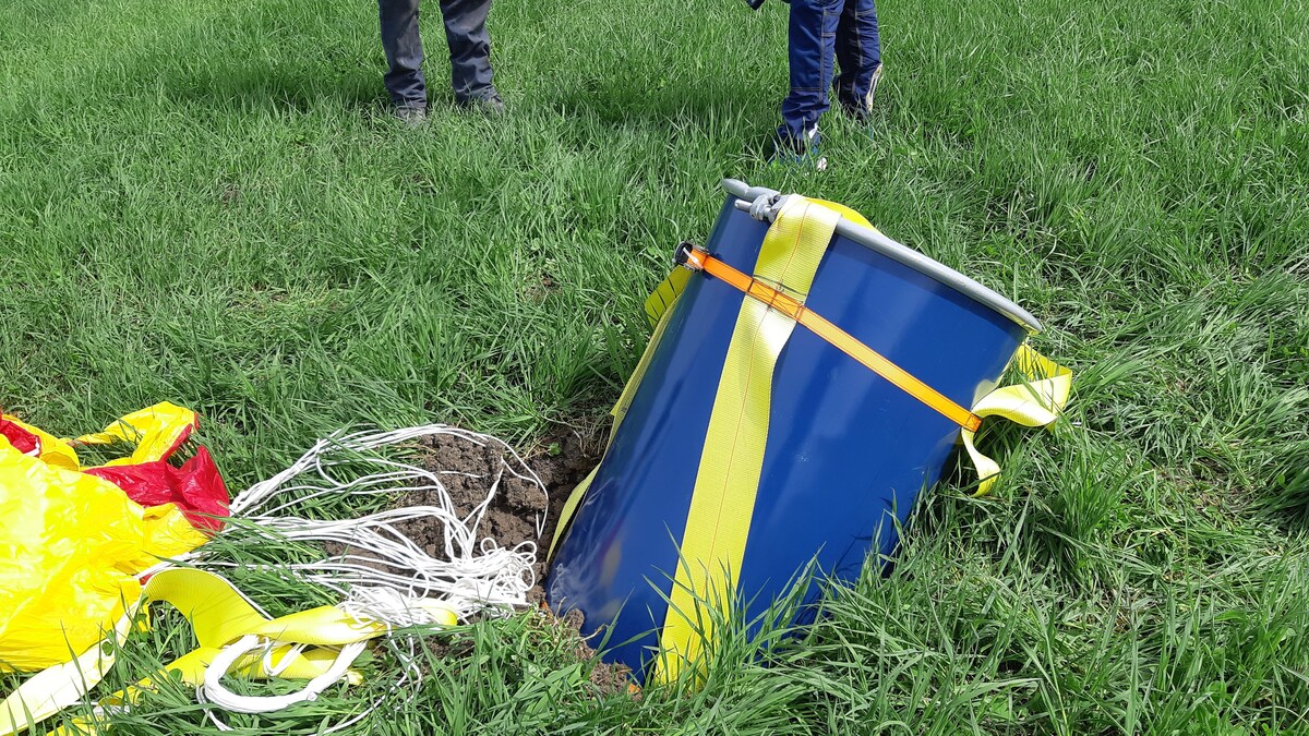 Un baril bleu attaché à un parachute jaune et rouge écrasé dans un champ.
