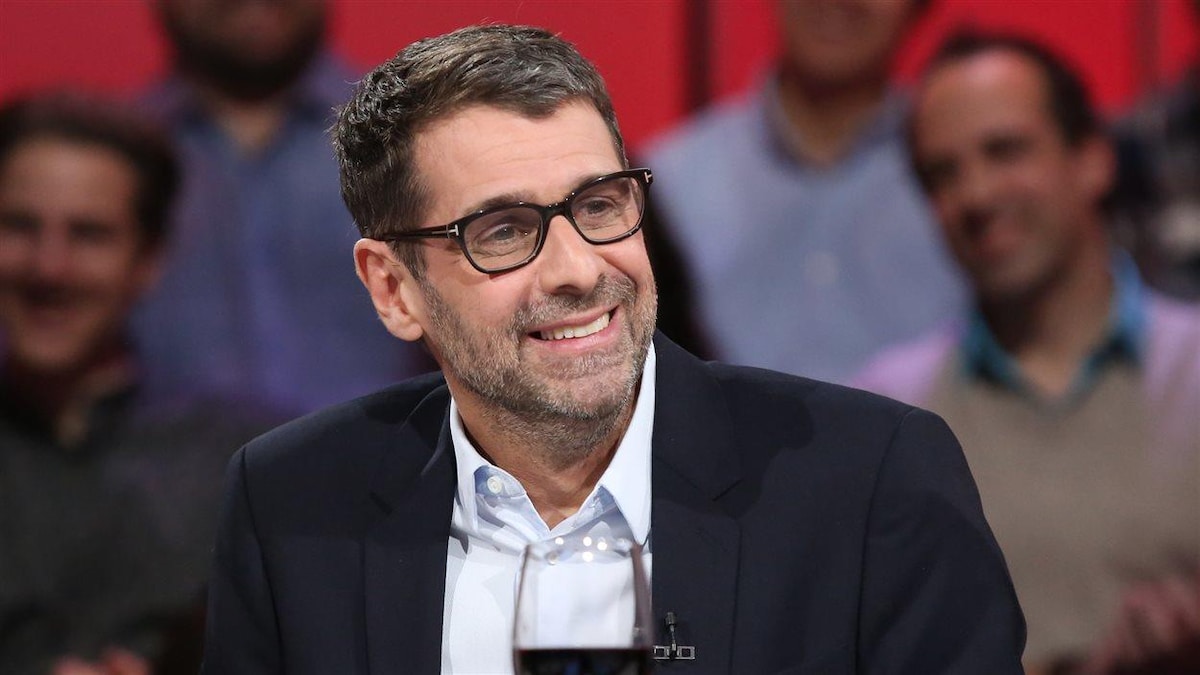 Un homme avec des lunettes et les cheveux grisonnants est assis devant uen foule, dans un studio de télévision.