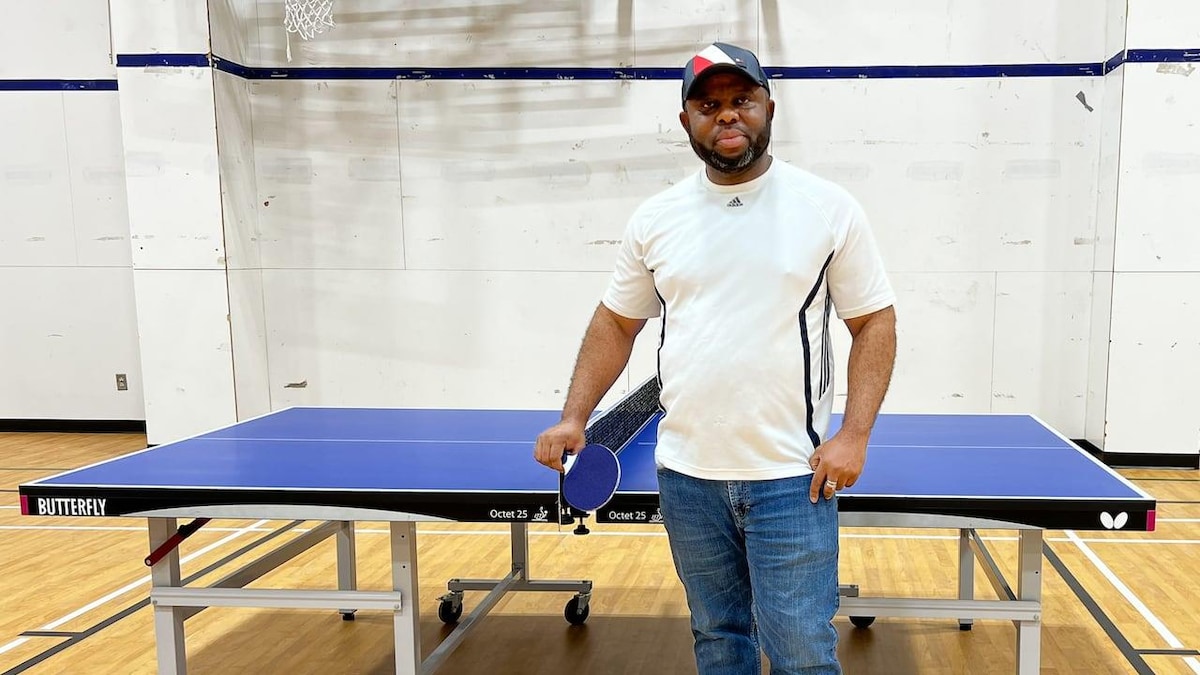 Le père Anyanwu pose devant une table de tennis de table avec une raquette dans la main.