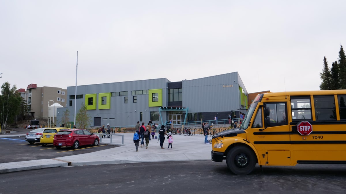 Un autobus est stationné devant le terrain d'une école, où se trouvent aussi quelques enfants et adultes, ainsi que trois voitures dans un stationnement.