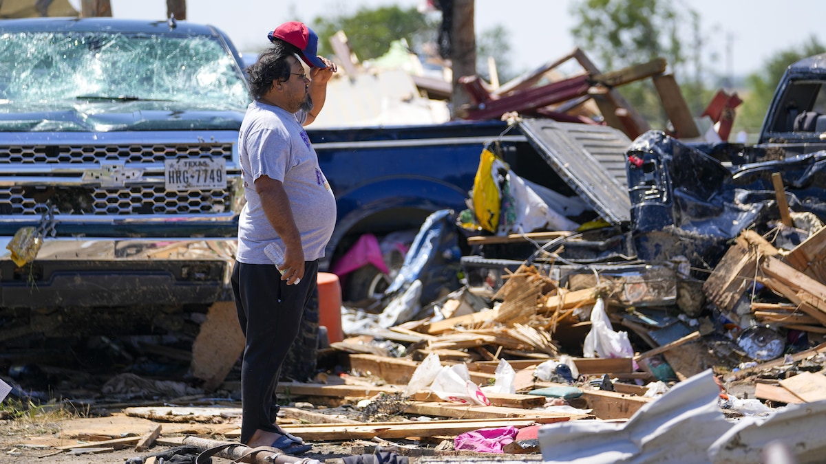 Debout dans des débris laissés par le passage d'une tornade, un homme soulève sa casquette pendant qu'il constate l'ampleur des dégâts.