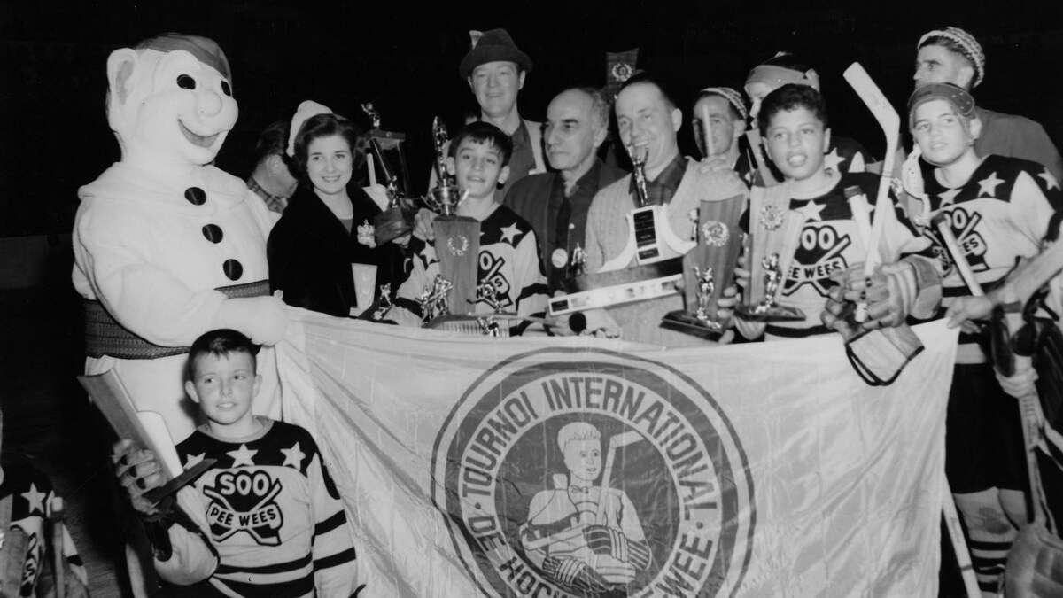 En 1963, le Bonhomme Carnaval, la reine du Carnaval, Lise Mercier, et des gagnants, hommes et garçons non identifiés, du tournoi international de hockey pee-wee qui montrent fièrement leurs trophées. Les garçons portent le chandail du tournoi. Devant le groupe est déployé le drapeau du tournoi.