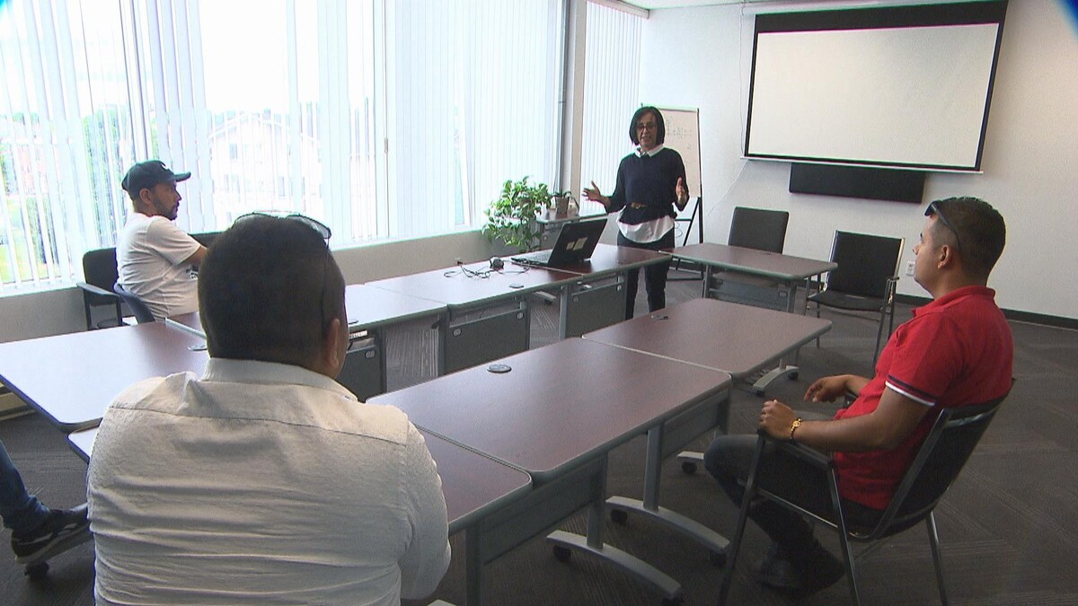 Trois hommes sont assis dans une salle de réunion. Ils regardent une femme debout qui fait une présentation. 