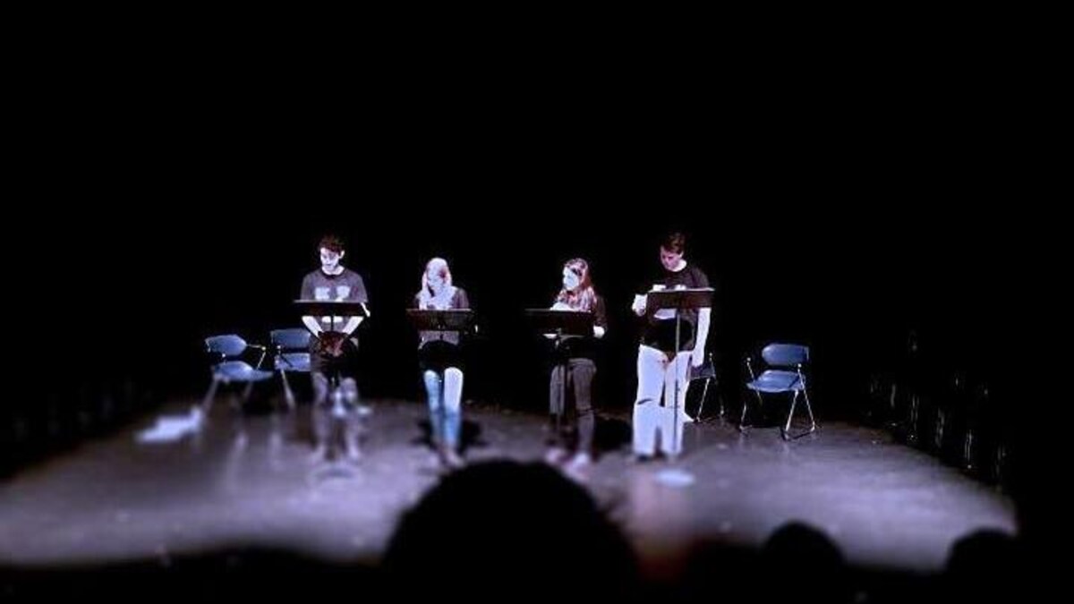 Sur une scène, quatre acteurs sont devant des lutrins pour la mise en lecture d'un texte.