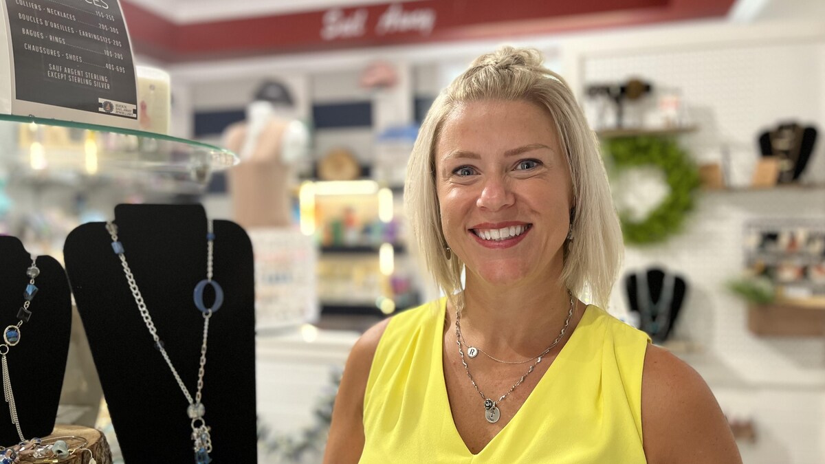 Femme blonde avec robe jaune sourit à la caméra près d'un étalage de bijoux.