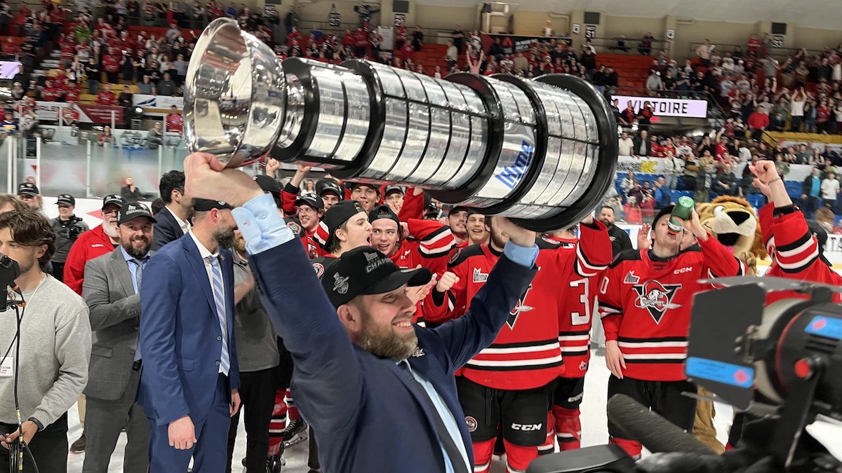 Un homme portant un complet et une casquette de champions soulevant un trophée au bout de ses bras devant des joueurs de hockey.
