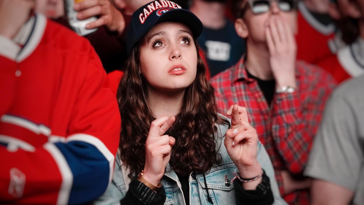Une jeune femme avec une casquette du Canadien a les doigts croisés et le regard attentif, pointé vers un écran géant à l'extérieur lors d'un rassemblement. 