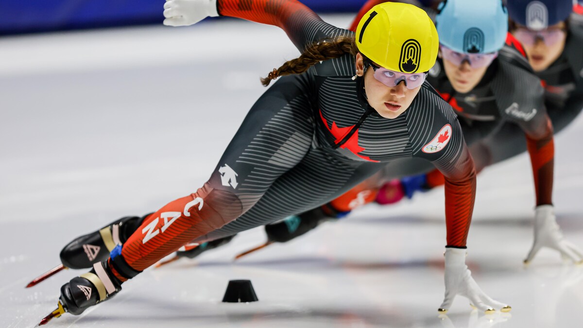 Une patineuse de vitesse, vêtue d'une combinaison rouge et noire, pose la main sur la glace en faisant un virage. 