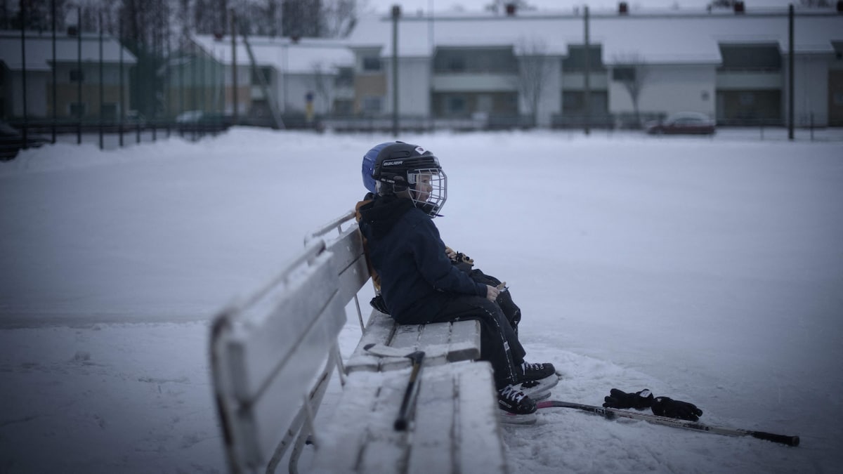 Un enfant portant un casque de hockey est assis sur un banc, devant ses gants et son bâton posés au sol.