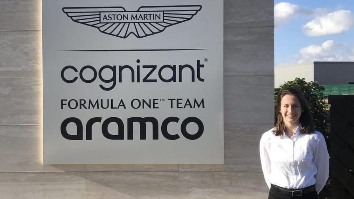 Une jeune femme est debout, dehors, à côté d'un grand panneau à l'entrée de l'usine d'une équipe de F1.