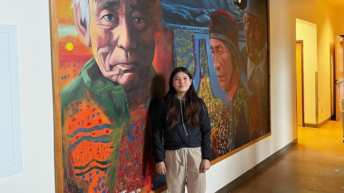 Mika Tenegan devant une murale située dans le hall de l'école de Pakuashipi. L'œuvre représente son arrière-grand-père, Shimun Mestenapeo.