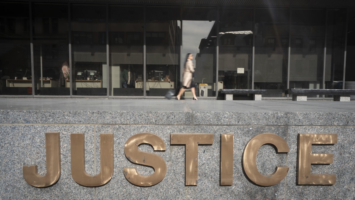 Une femme tirant une petite valise sur roulettes marche devant le palais de justice de Montréal avec à l'avant-plan le logo « Justice ».
