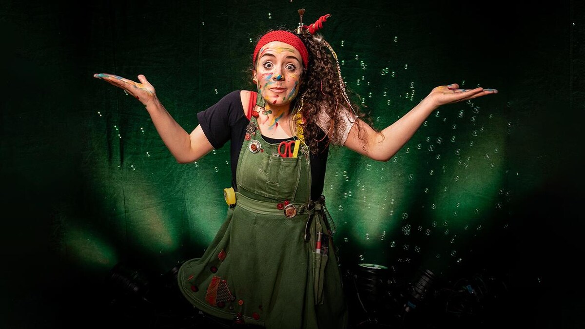 Ariane DesLions, vêtues d'une salopette verte, le visage barbouillé de peinture de diverses couleurs, entourée de bulles de savon, les bras de chaque côté d'elle, comme si elle se posait une question. L'éclairage derrière elle est vert.
