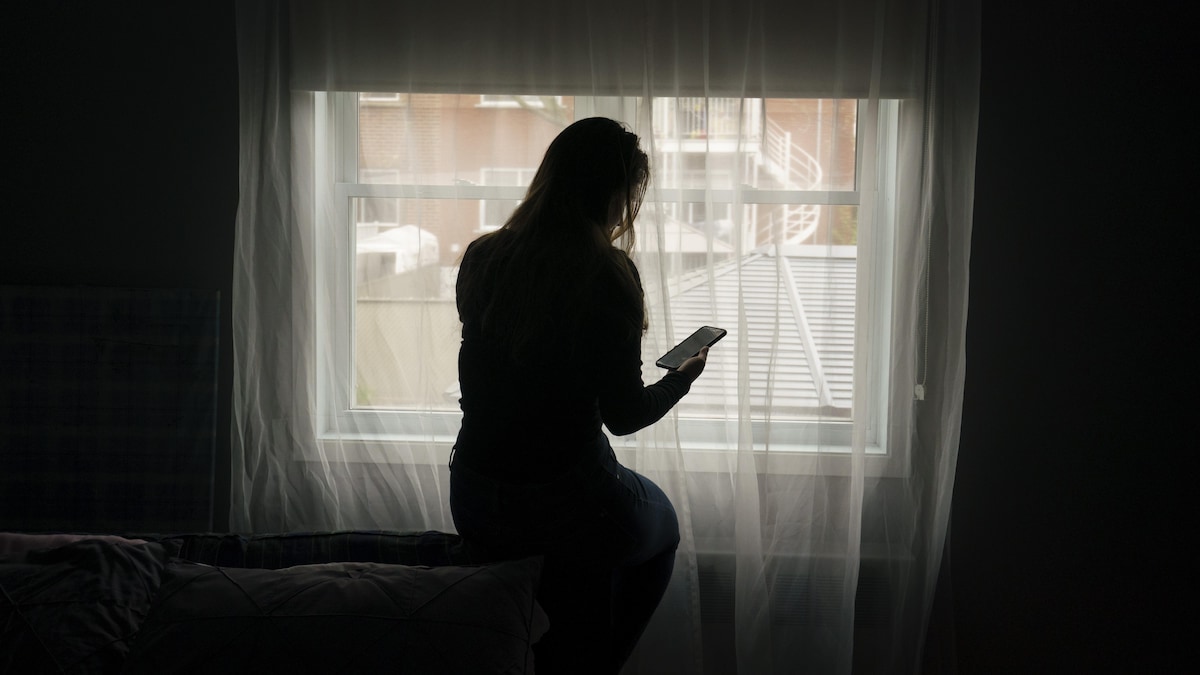 Une femme consulte son téléphone cellulaire assise dans une pièce plongée dans la pénombre, devant une fenêtre d'un quartier résidentiel.