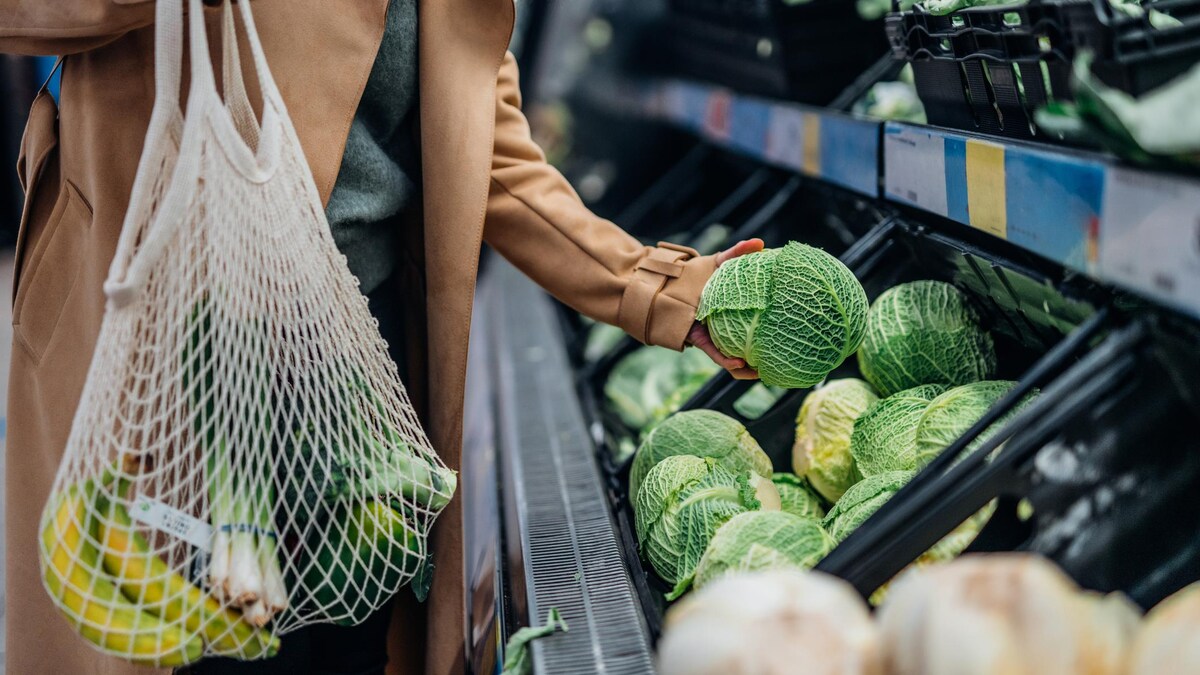 Photo recadrée d'une femme faisant des emplettes dans un supermarché. Elle s'apprête à mettre un chou dans un sac à provisions en filet de coton. 