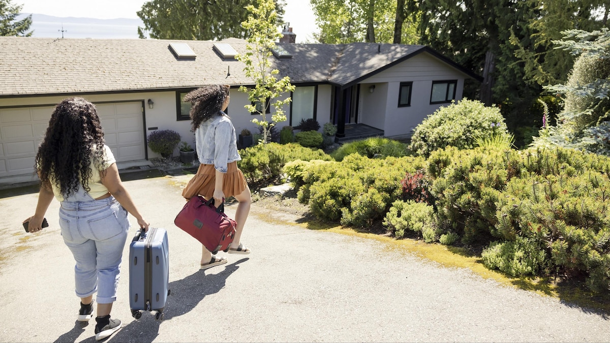 Deux jeunes femmes marchent avec leurs bagages vers l'entrée d'une maison lors d'une journée ensoleillée.