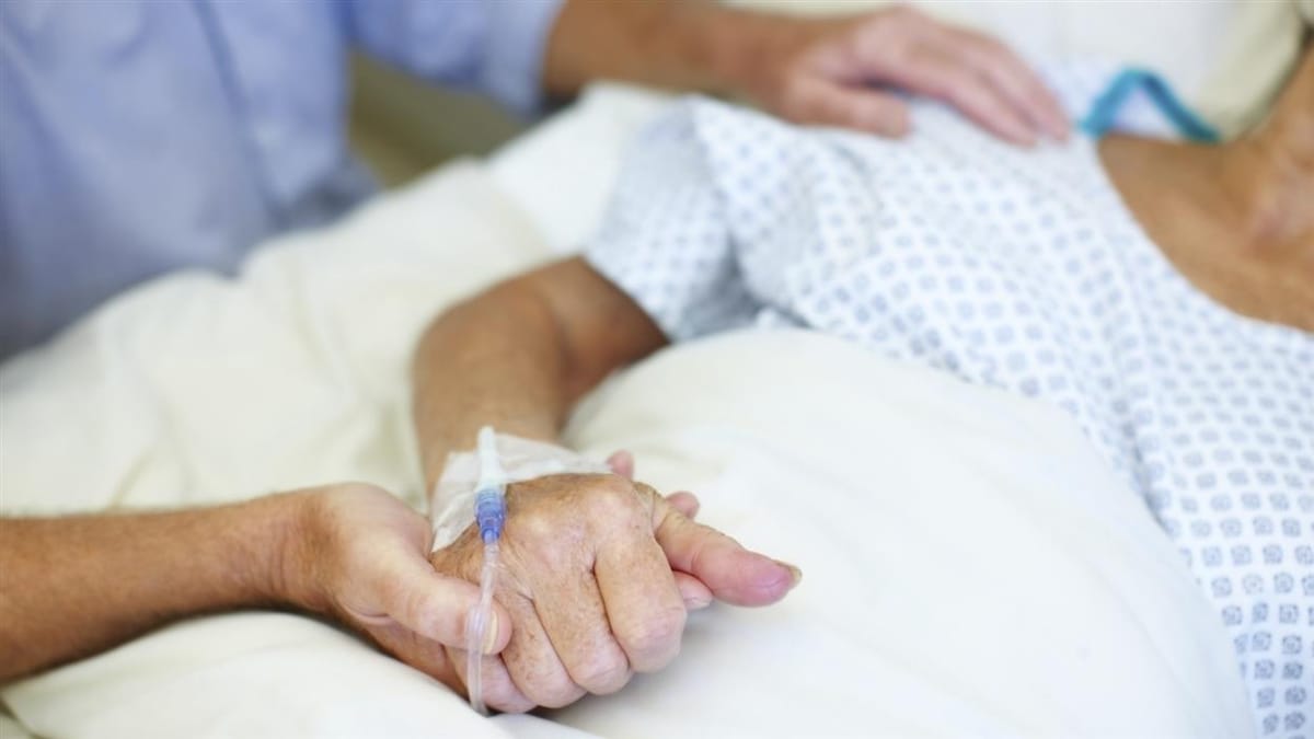 Gros plan sur une personne alitée aux soins palliatifs. Un individu la tient par la main. 