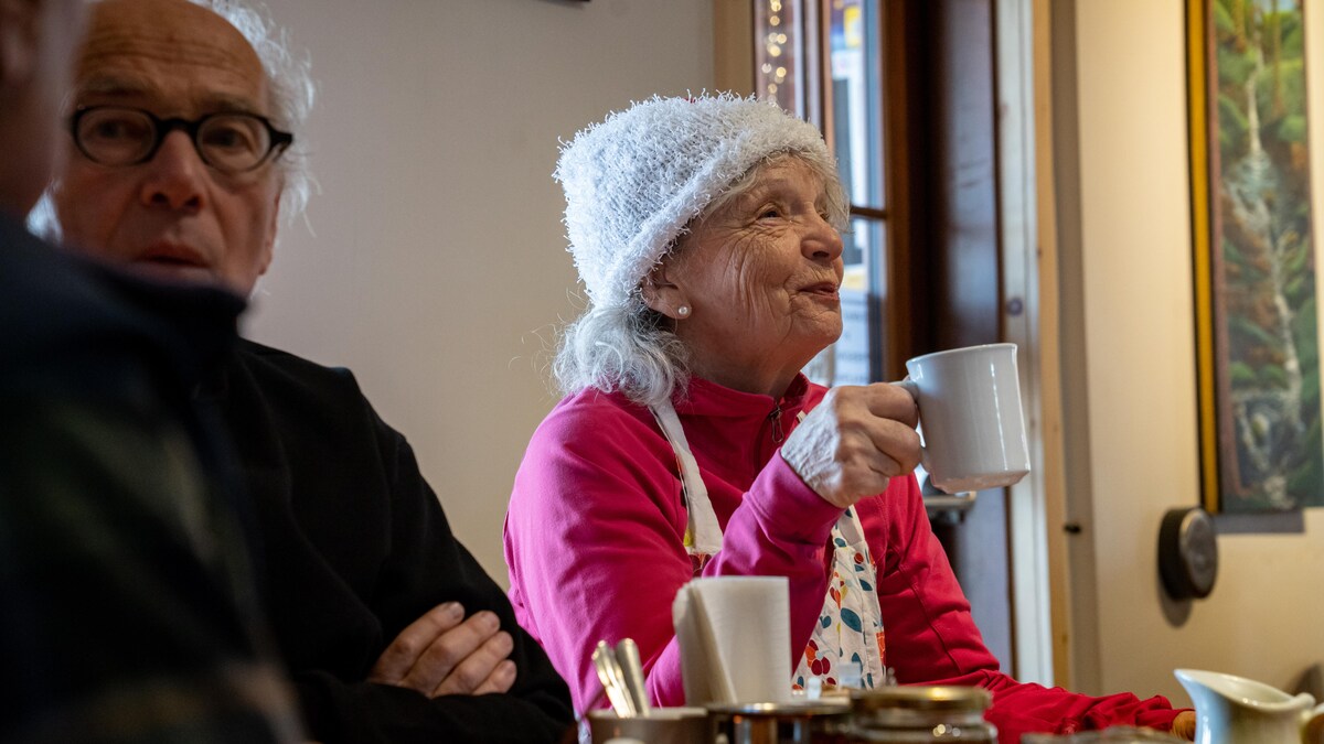 Deux aînés à la table d'un restaurant. À gauche un homme parle avec son voisin (qui n'est pas sur la photo). À droite, une femme sourit, une tasse de café à la main. 