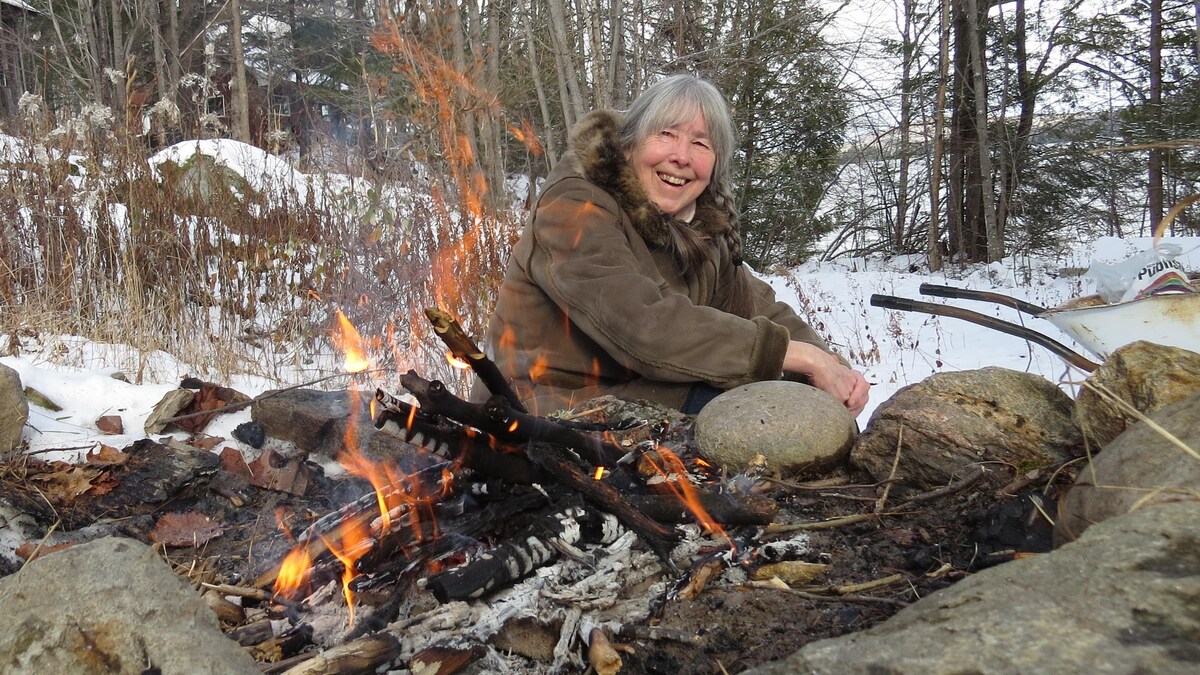Francine Ouellette, souriante, assise au bord d'un feu dans une forêt.