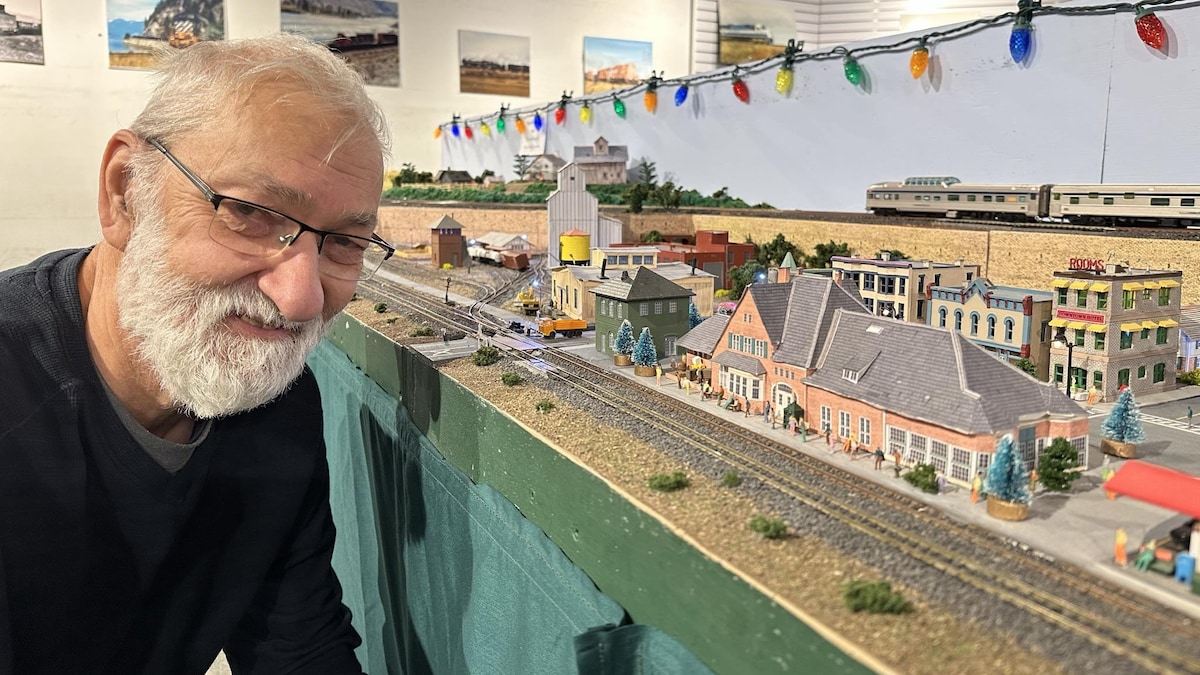 Gilles Doiron est devant une partie de l'exposition de train. On voit des édificies miniature derrière lui. 