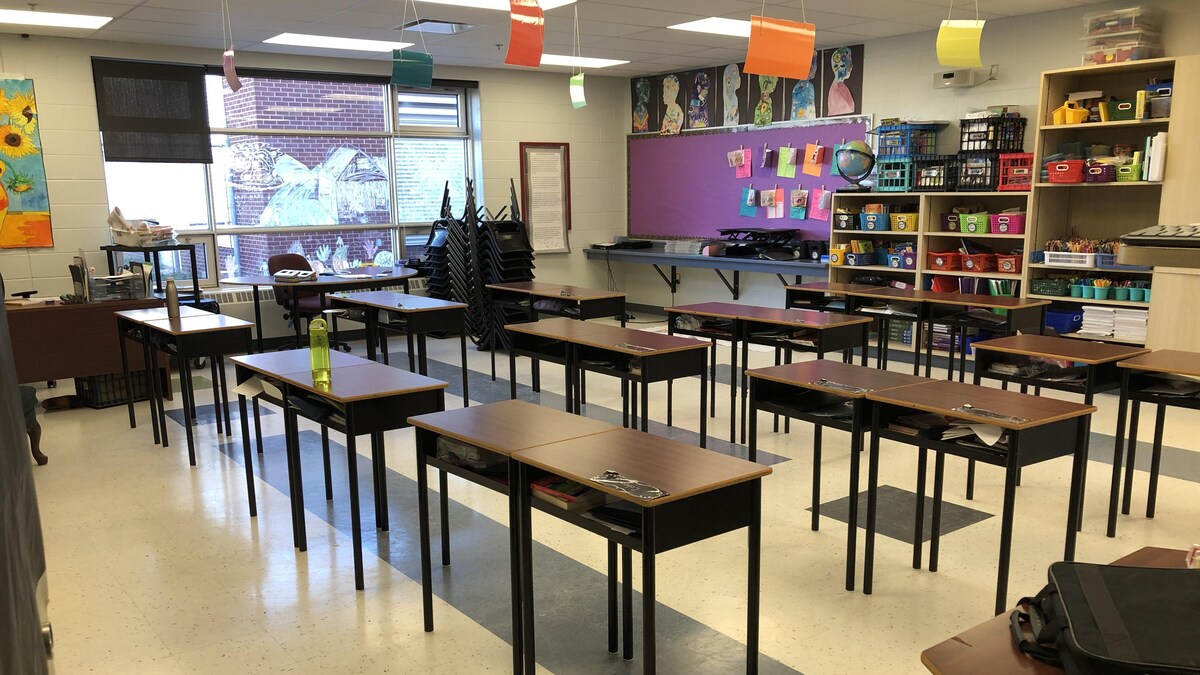 Une salle de cours avec dix-neuf bureaux aménagés au centre. Des fournitures scolaires sont déposées sur des étagères au fond. 