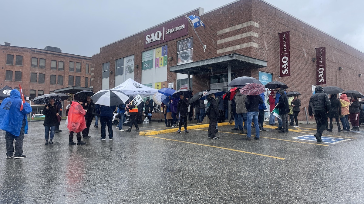 Des gens avec des parapluies manifestent devant une succursale de la Société des alcools du Québec.