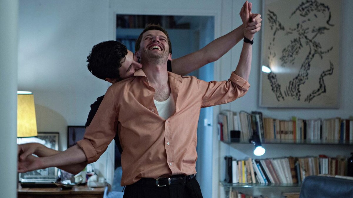 Dans un salon, deux hommes souriants dansent ensemble, l'un embrasse l'autre dans le cou.