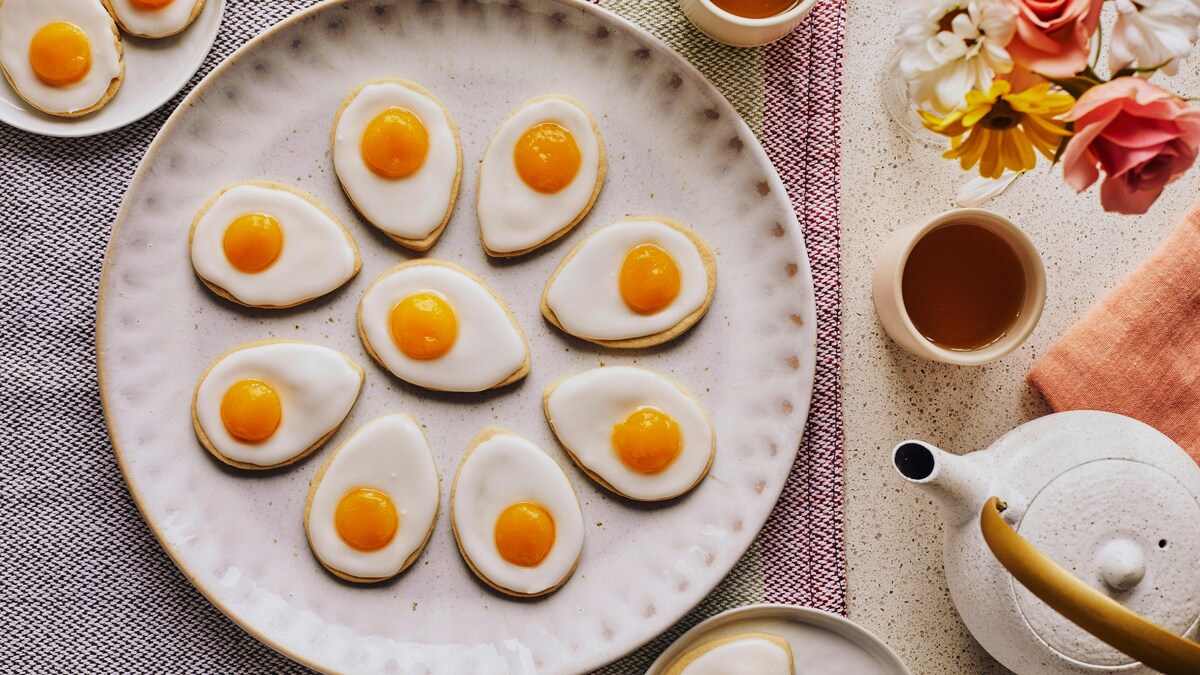 Dans une grande assiette de service, il y a plusieurs biscuits en forme d’œufs, avec une théière, des tasses de thé et des fleurs.