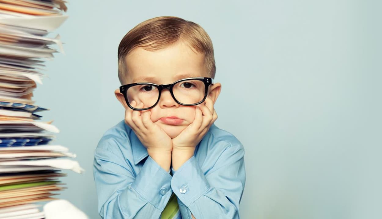 Un garçon, portant des lunettes et une cravate, appuie sa tête sur des mains, assis à côté d'une pile de documents.