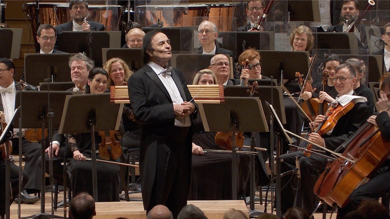 Charles Dutoit est debout sur scène, au milieu d'un orchestre. Charles Dutoit est debout sur scène, au milieu d'un orchestre.