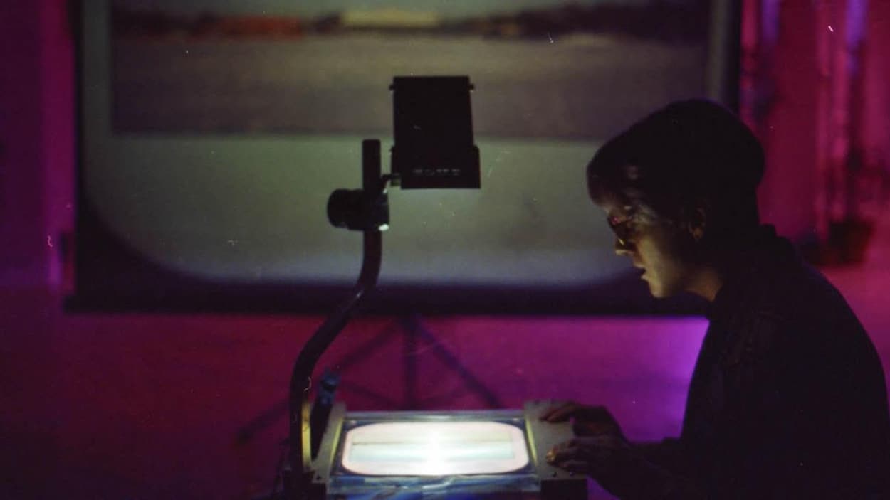 Une personne portant des lunettes et vue de profil regarde une image posée sur un rétroprojecteur. L'image floue d'un paysage est projetée sur un écran sur trépied, en arrière-plan.