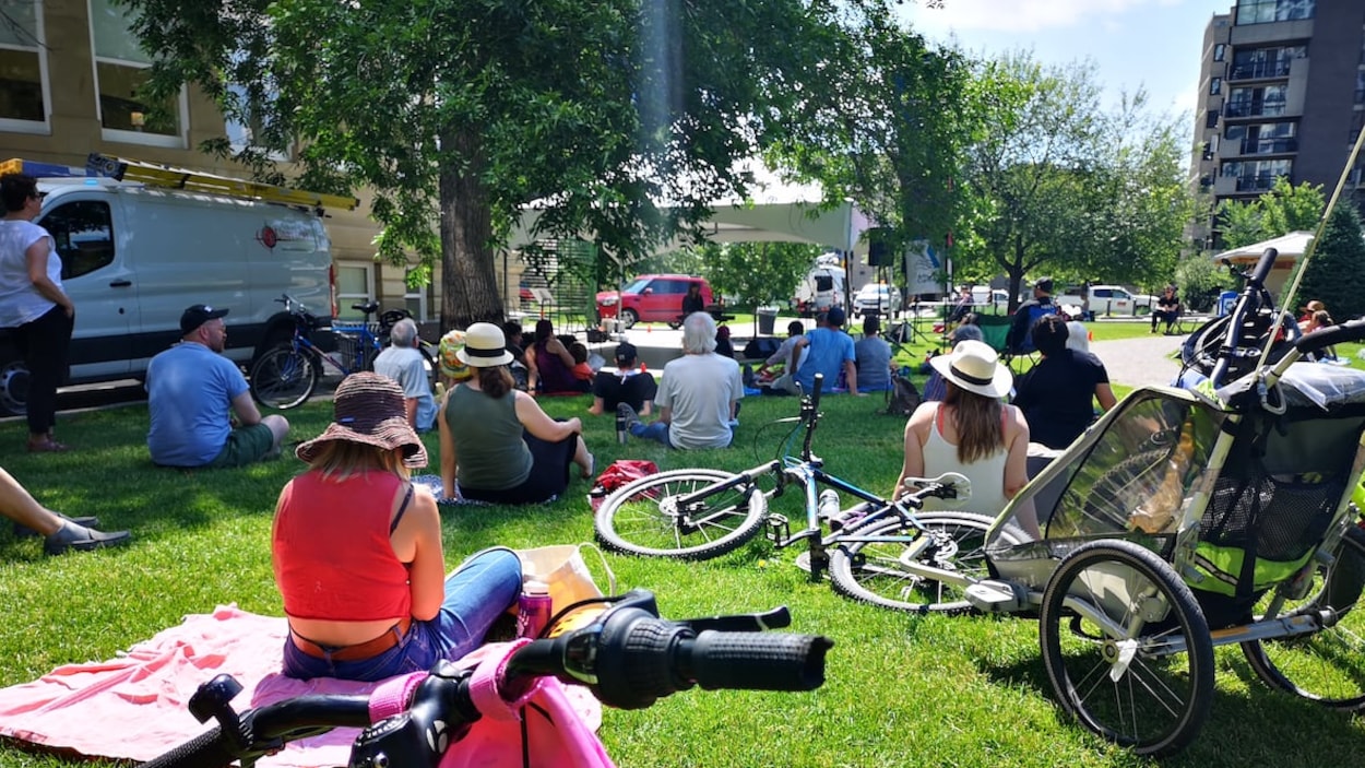 Des gens assis sur l'herbe, dans un parc, regardent vers une scène dressée sous une tente.