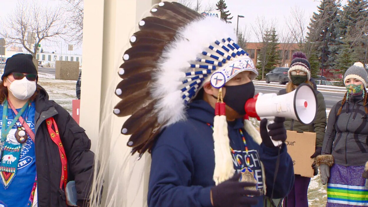 Le chef Billy Morin, portant une coiffe de plumes, s'adresse à la foule rassemblée sur le trottoir en parlant dans un mégaphone.