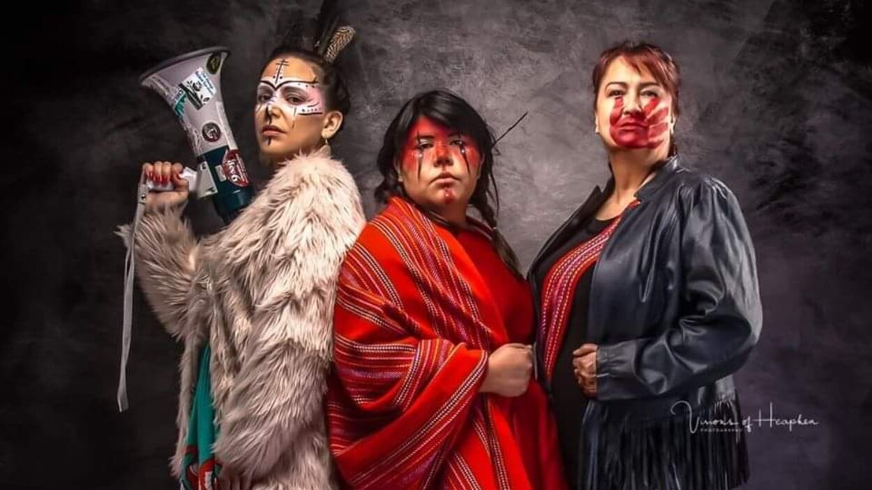 Trois femmes en habits traditionnels et portant du maquillage posent pour l'oeuvre. L'une d'entre elles tient en main un porte-voix.