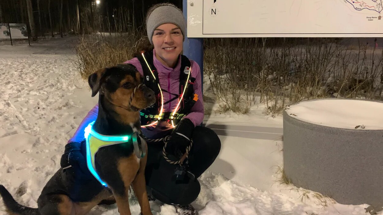 Jessica Hoskins pose, accroupie à côté de son chien, sur un sentier, un soir d'hiver. Les deux portent des bandes lumineuses.