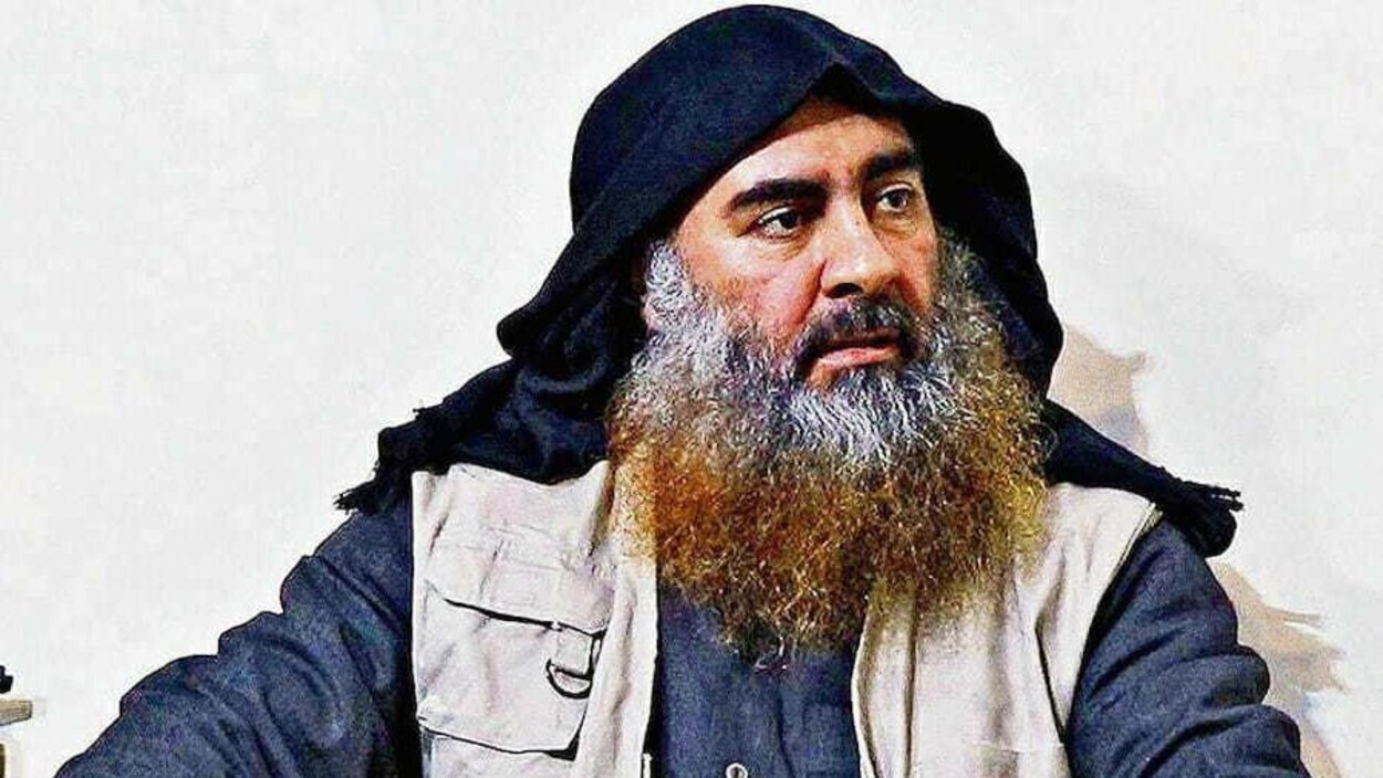 Abou Bakr al-Baghdadi, assis, porte un capuchon, une veste, une chemise et une barbe à deux couleurs (blanche et blonde).