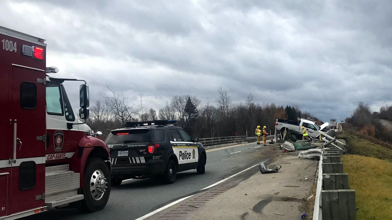 Photo de la scène d’accident. On aperçoit une camionnette blanche accidentée. Un véhicule utilitaire sport de la Sûreté du Québec et un camion de pompier. Photo de la scène d’accident. On aperçoit une camionnette blanche accidentée. Un véhicule utilitaire sport de la Sûreté du Québec et un camion de pompier.