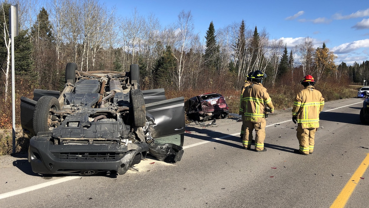 Trois pompiers discutent devant deux véhicules accidentés au bord d'une route.