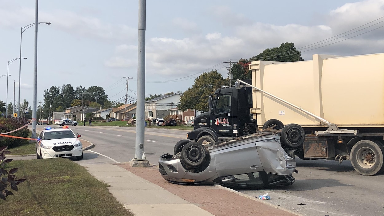 Une voiture compact renversée, dont la partie arrière est lourdement endommagée. Près de cette voiture sont stationnés un camion d'ordures et une voiture du Service de police de la Ville de Gatineau.
