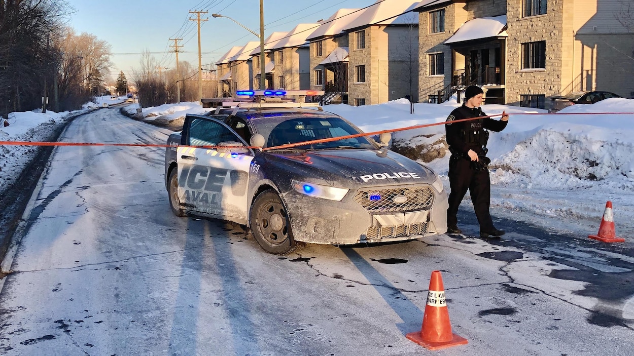 Une voiture de police immobilisée dans une rue, un policier s'apprête à traverser un cordon de sécurité.