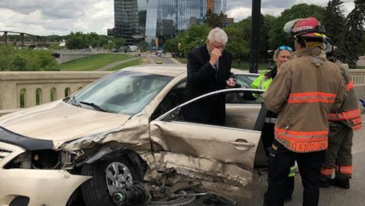 Un passager et des premiers répondants debout devant la voiture endommagée par l'accident.