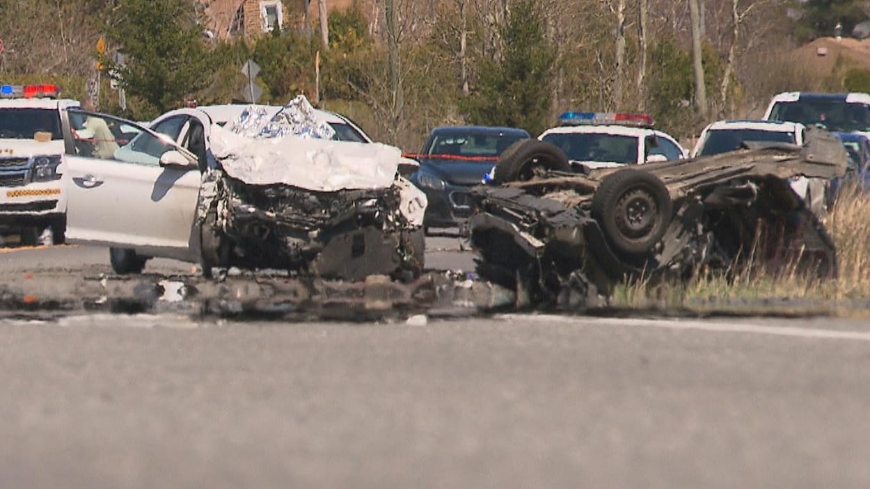 Un voiture noire est sectionnée et se trouve sur le toit. Une autre voiture est abîmée à l'avant. En arrière, il y a des voitures de police. Un voiture noire est sectionnée et se trouve sur le toit. Une autre voiture est abîmée à l'avant. En arrière, il y a des voitures de police.