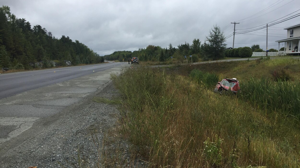 Une voiture rouge est partiellement cachée par de l'herbe dans un ravin en bordure de route et une voiture de police est stationnée loin devant.