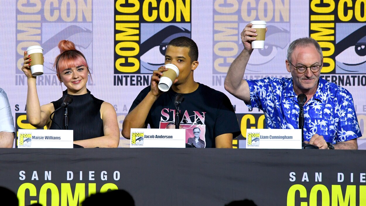 Les trois acteurs sont assis derrière une table identifiée du logo du Comic-Con de San Diego et lèvent chacun une tasse de café jetable.
