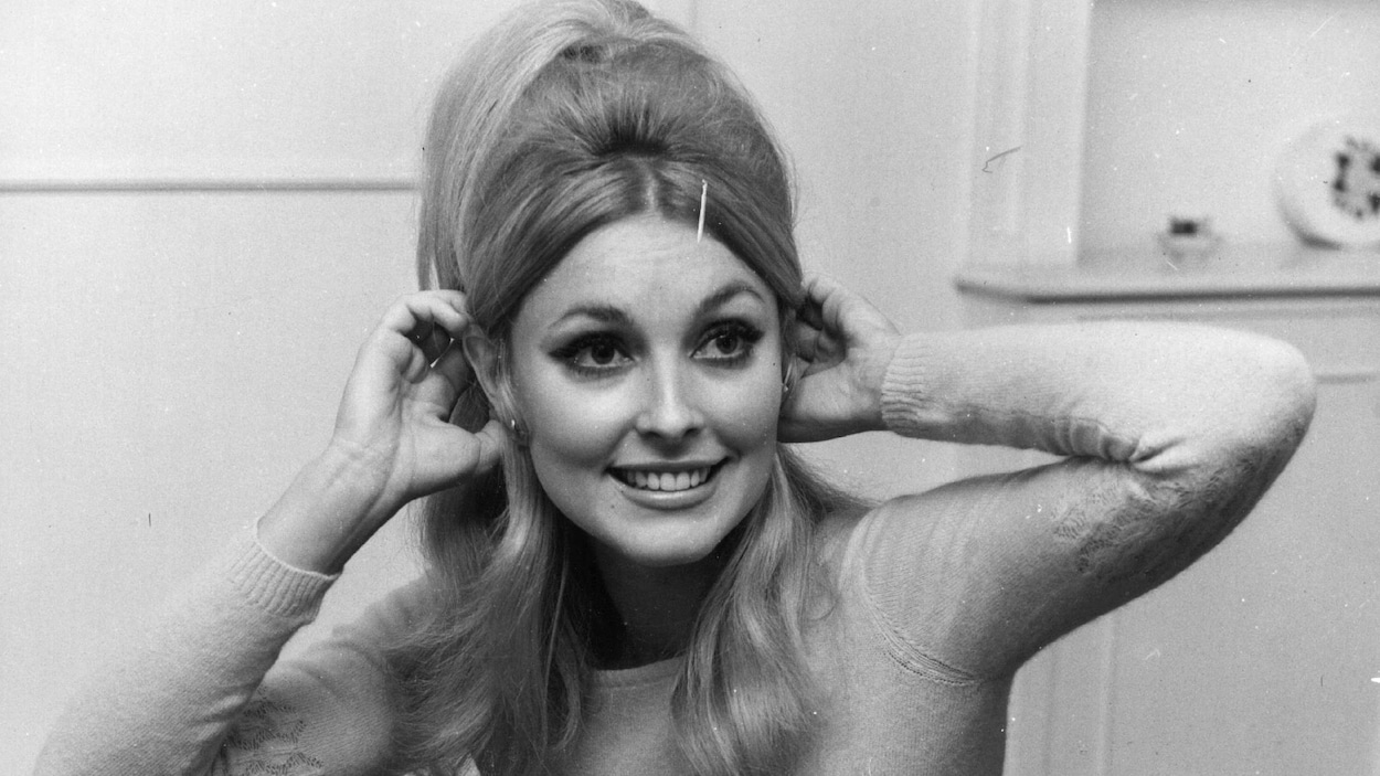 La jeune femme touche ses oreilles avec ses mains et sourit dans une photo en noir et blanc.