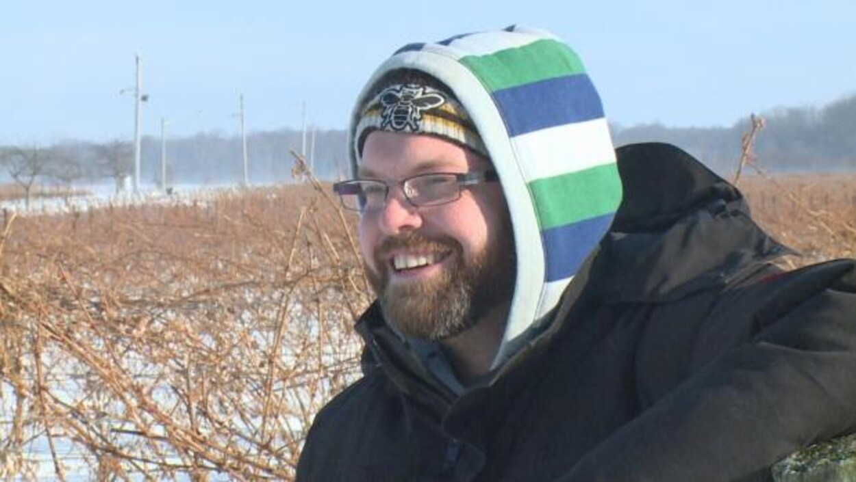 Adam Graham dans un champ. Il sourit et porte des lunettes et une tuque, il a aussi une barbe. Adam Graham dans un champ. Il sourit et porte des lunettes et une tuque, il a aussi une barbe.
