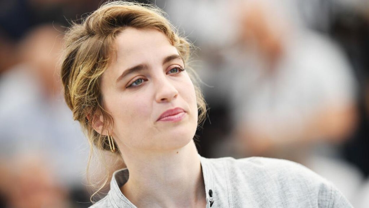 Elle porte une tenue grise et ses cheveux blonds sont relevés.