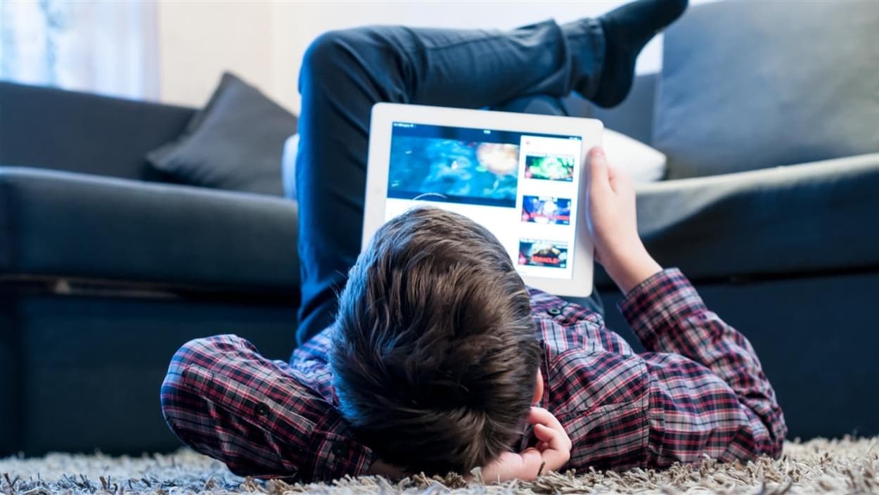 Un adolescent regarde une tablette allongé sur le sol du salon. 