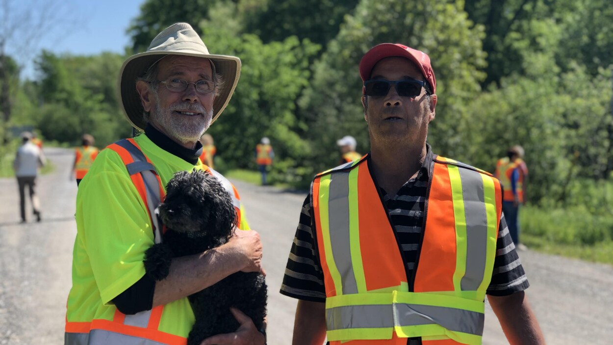 Deux hommes vêtus d'un dossard posent, l'un d'eux tient un petit caniche dans les bras
