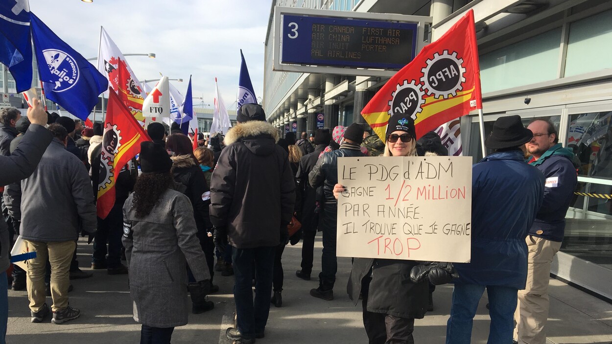 Des gens manifestent devant l'aéroport de Montréal, une femme brandissant une pancarte sur laquelle il est écrit: «Le PDG d'ADM gagne un demi-million par année. Il trouve que je gagne trop.»